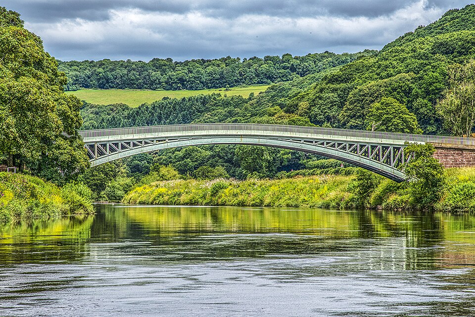 Bigsweir Bridge from the river bank in summer: a single pale green iron arch springing across the full width of the Wye, the river calm and reflective below, with steep wooded hills and one bright field rising behind it.