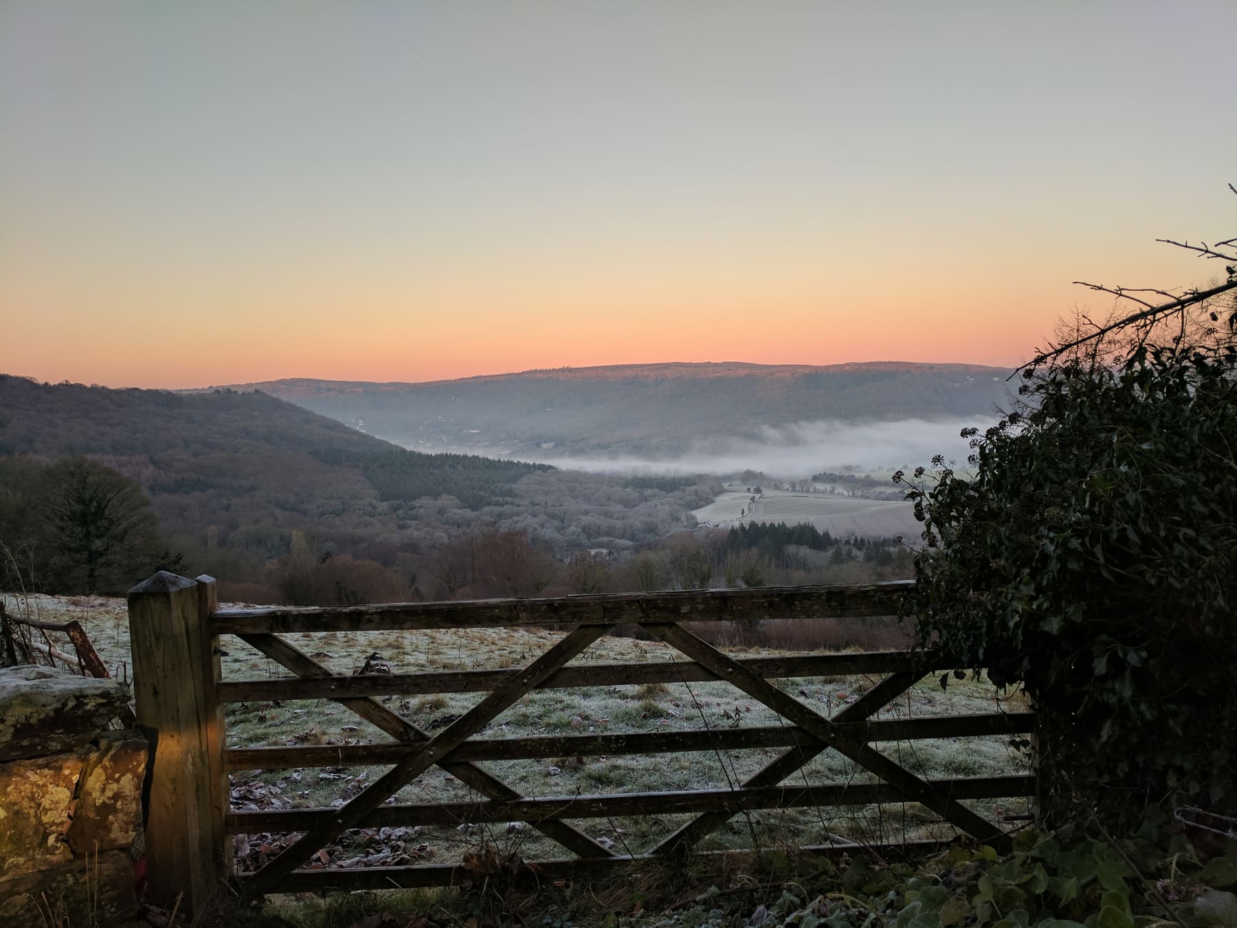 The gate topped with frost on a January dawn, the field white behind it, mist lying along the valley floor, and the sky glowing orange to pale gold above the dark line of the far ridge.