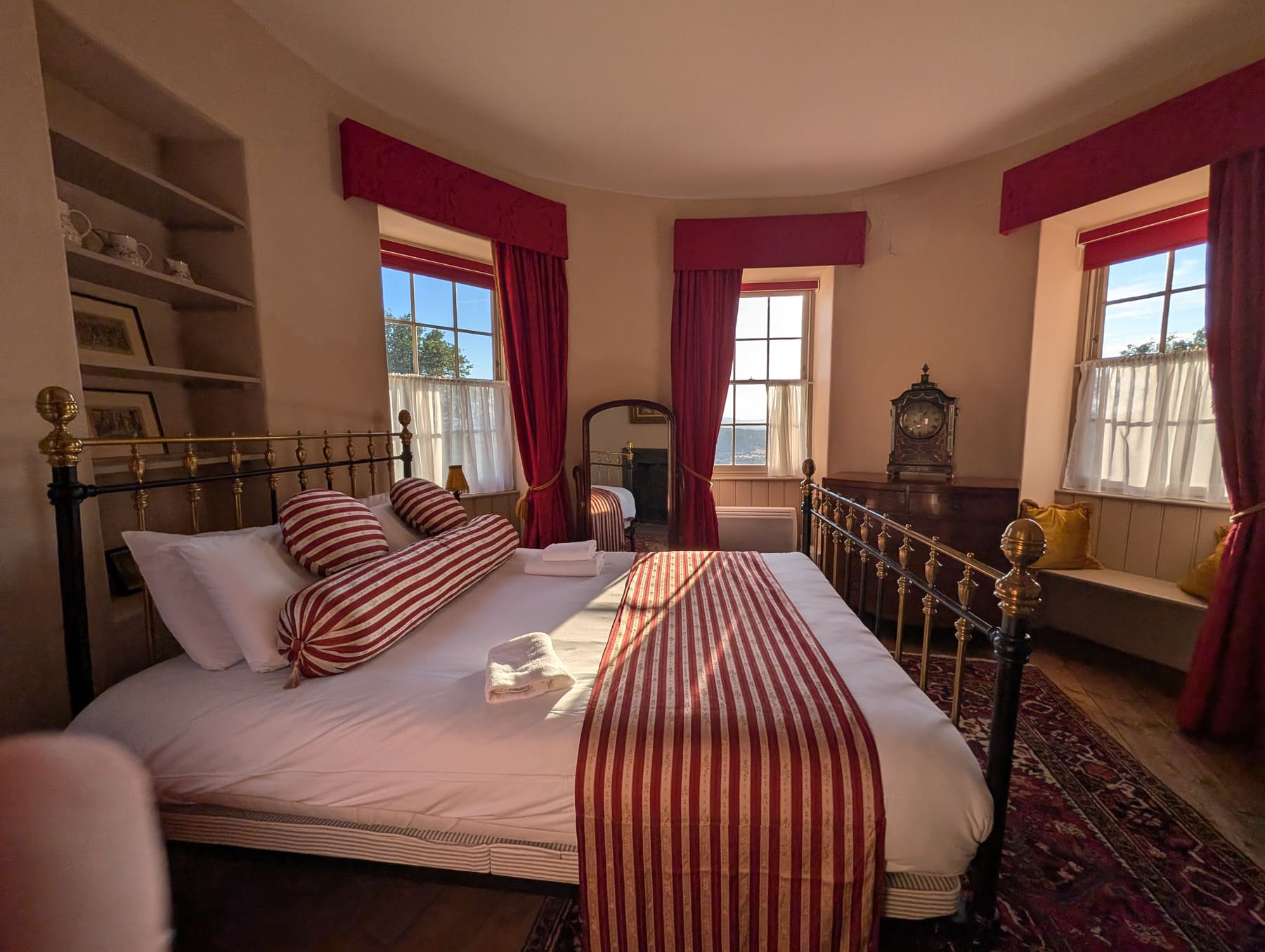 The round bedroom of the Kymin Round House: a brass and iron bed with red and white striped bolsters and runner, red curtains at three small sash windows, an antique clock on a chest of drawers, and low evening sun falling across the white bedspread.