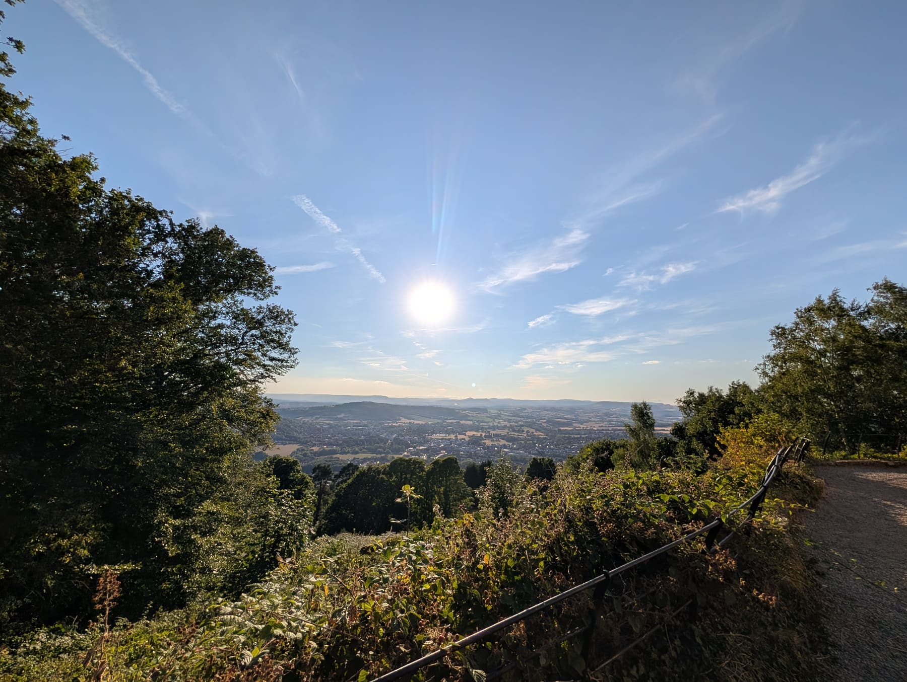 The gravel path curving around the top of the Kymin on a summer evening, the sun still high over hazy blue hills, Monmouth spread across the valley far below, with brambles and a black iron railing beside the path.