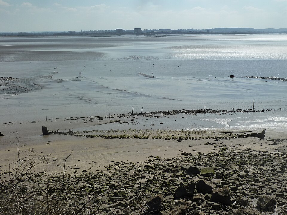 The ribs of a wrecked wooden boat lying half buried in grey tidal mud on the Lydney foreshore at low water, with more broken timbers and posts behind it, a bank of quarried stone rubble in the foreground, and the wide flat Severn beyond with the low blocks of Berkeley power station on the far shore.