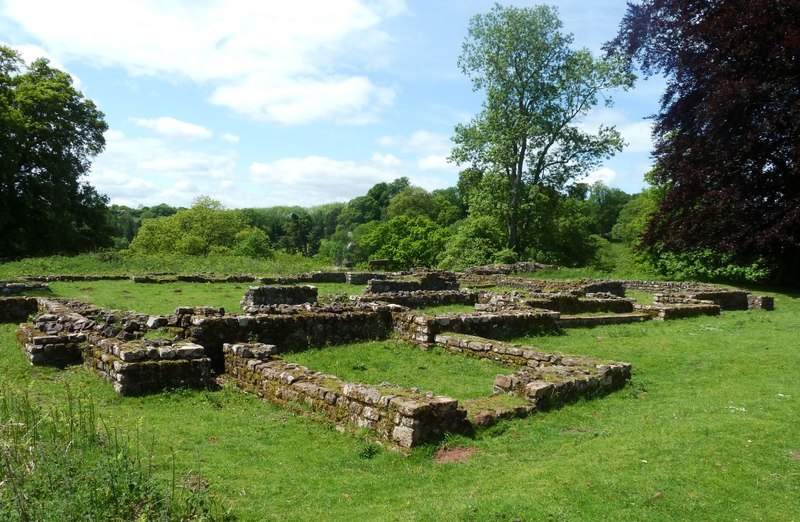 The excavated ruins of the Roman temple at Lydney Park on a sunny day: low mossy stone walls, perhaps knee high, laying out a rectangle divided into rooms and corridors, with mown grass growing inside and over them, and mature parkland trees and a copper beech behind.