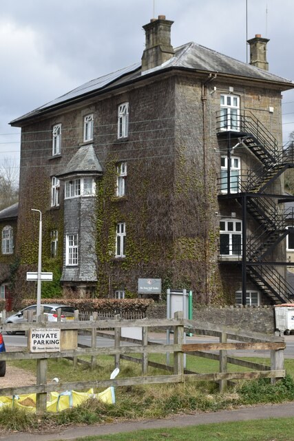 The old Parkend Ironworks blowing engine house: a four storey stone building almost entirely covered in ivy, with two tall chimneys, sash windows showing through the leaves, a slate hipped roof, black iron fire escape stairs zigzagging down one side, and a wooden fence with a private parking sign in front.
