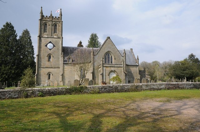 St Paul's church, Parkend, from across the grass: a stone church with a battlemented clock tower flying a flag, gabled arms reaching out from its centre, dark yews to the left, gravestones behind a low stone churchyard wall, under a bright spring sky.