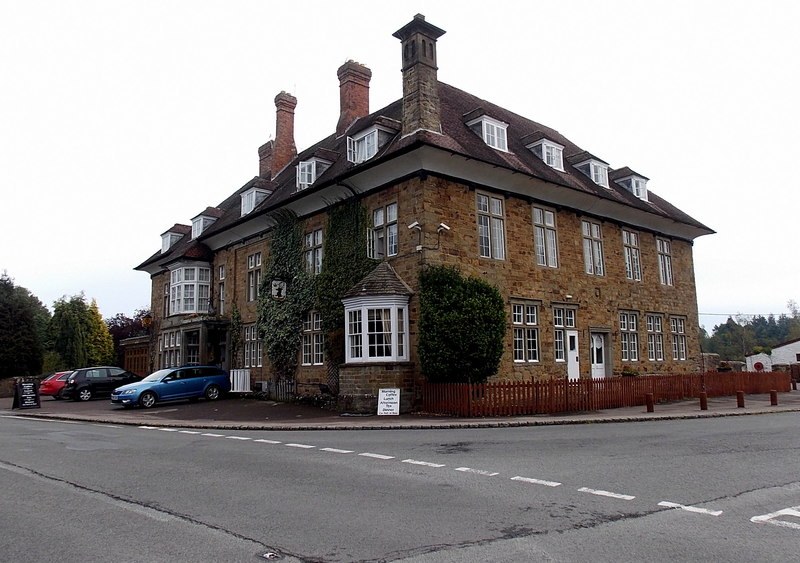 The Speech House Hotel from the road junction: a large seventeenth century stone building with tall brick chimneys, rows of white sash windows, dormer windows in a hipped roof, ivy on one corner, a small bay-windowed porch, and cars parked along the frontage under a grey sky.