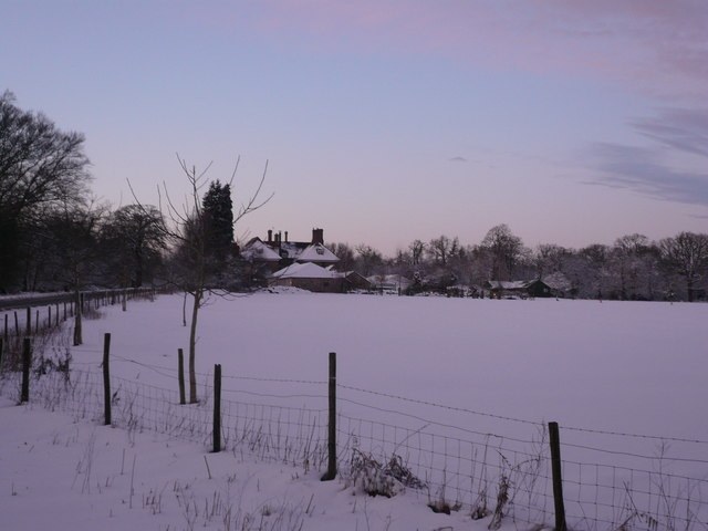 The Speech House seen across a snow-covered field at dusk: a distant stone house with clustered chimneys among bare winter trees, a line of young trees and a wire fence crossing the white field in the foreground, under a pink and violet sky.