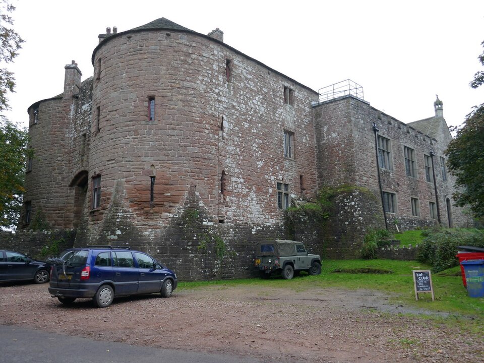 St Briavels Castle from its gravel forecourt: the two great round drum towers of the Edward I gatehouse in weathered pink and grey stone, with the long hall range behind, and parked in front of it the everyday cars and Land Rover of the youth hostel the castle now is, with a wheelie bin by the wall.