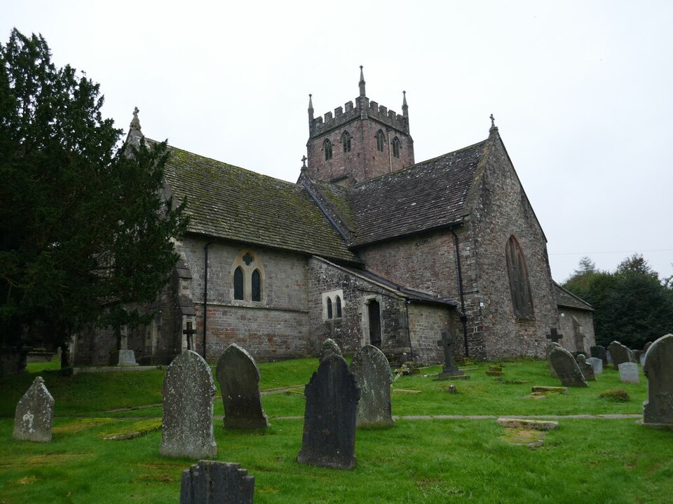 St Mary's church at St Briavels under a plain grey sky: a Norman cruciform church of mixed pink and grey stone with a squat battlemented central tower, mossy roofs at several angles, and leaning headstones in the grass of the churchyard.