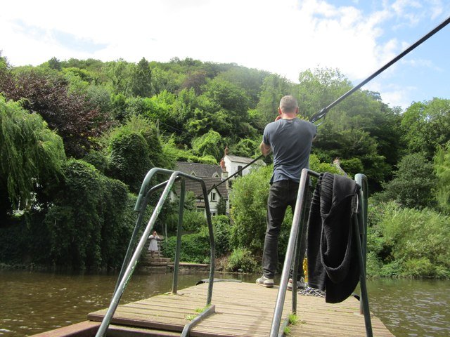 The hand ferry at Symonds Yat in high summer: a ferryman in a grey T-shirt stands on the wooden deck of the small flat ferry, hauling it across the green River Wye on an overhead cable, with the steep wooded gorge rising in full leaf behind him and a stone cottage half hidden in the trees on the far bank.